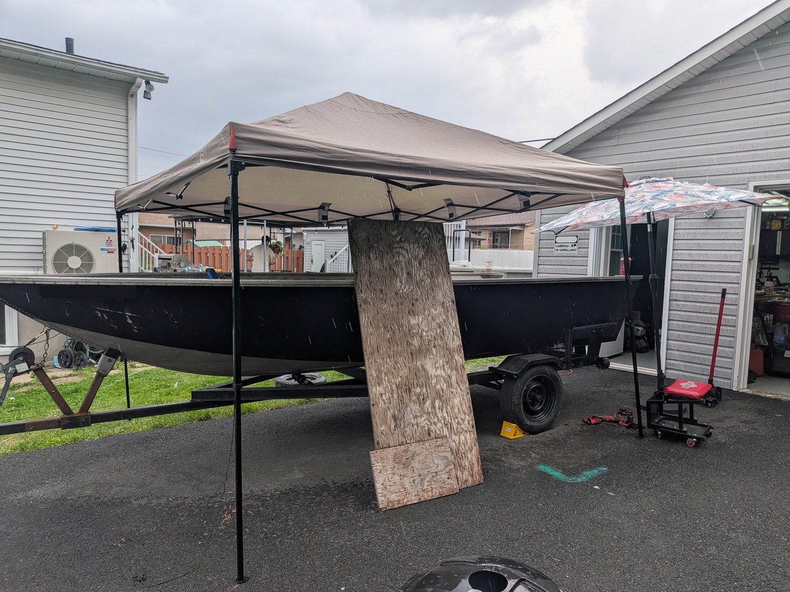 Le bateau sous un abri de toile dans l'entrée, un jour de pluie, une grande feuille de contreplaqué appuyée contre le montant : le chantier se fait dehors.