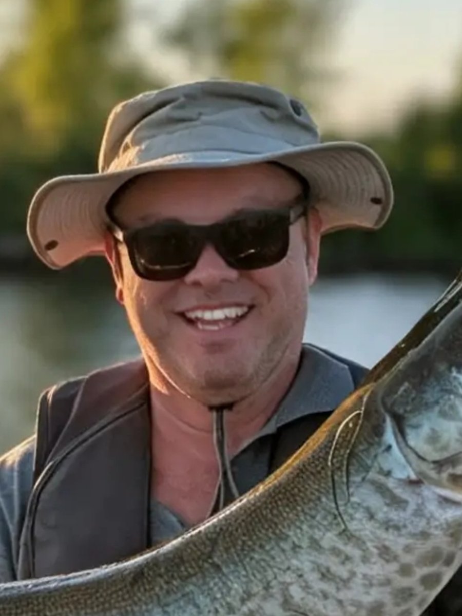 BOBE en gros plan au coucher du soleil, chapeau de brousse et lunettes fumées, la tête d'un maskinongé à côté de lui