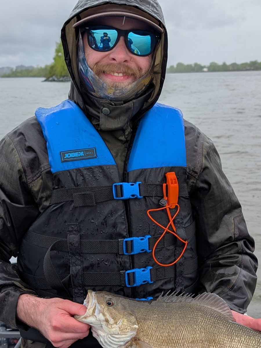 Kevin Caron en gros plan sur l'eau par temps gris, capuchon relevé, lunettes fumées et gilet de sauvetage, tenant sa prise